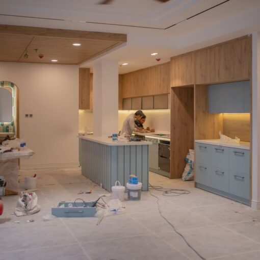 Under-construction kitchen with a blue island, wooden cabinets, and a worker measuring at the counter.