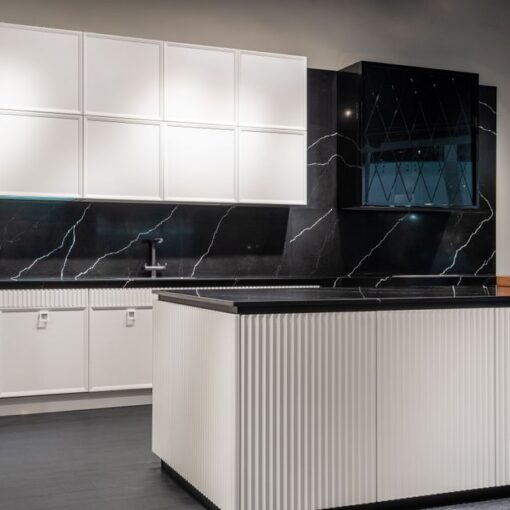 Modern white and black kitchen display: white upper and lower cabinets, black marble backsplash, and a grooved white island in a showroom.