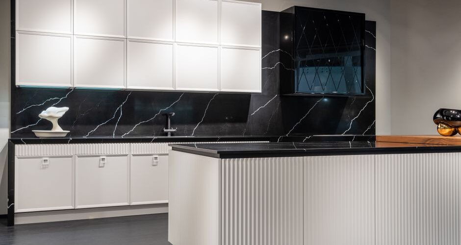 Modern white and black kitchen display: white upper and lower cabinets, black marble backsplash, and a grooved white island in a showroom.