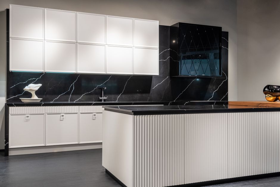 Modern white and black kitchen display: white upper and lower cabinets, black marble backsplash, and a grooved white island in a showroom.