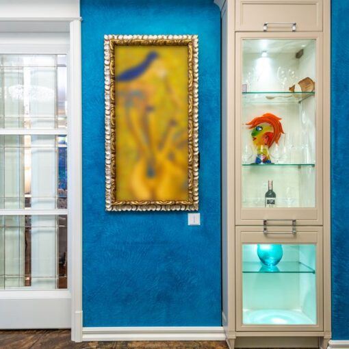 Blue textured wall with a gold-framed blurred abstract painting, placed next to a beige glass-front cabinet displaying a colorful fish-head sculpture, a bottle, and a blue glass orb on the lower shelf across from white doors.