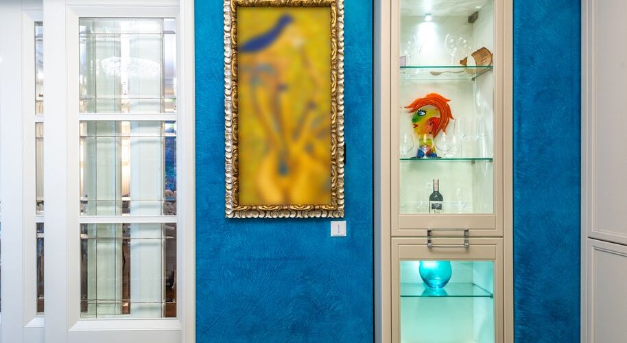 Blue textured wall with a gold-framed blurred abstract painting, placed next to a beige glass-front cabinet displaying a colorful fish-head sculpture, a bottle, and a blue glass orb on the lower shelf across from white doors.