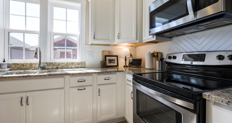 Bright, modern kitchen with white cabinets, granite counters, and stainless steel stove and microwave; double sink by windows on the left.