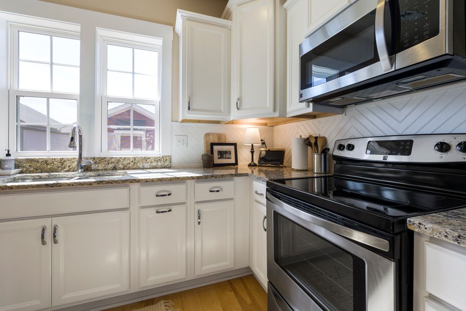 Bright, modern kitchen with white cabinets, granite counters, and stainless steel stove and microwave; double sink by windows on the left.