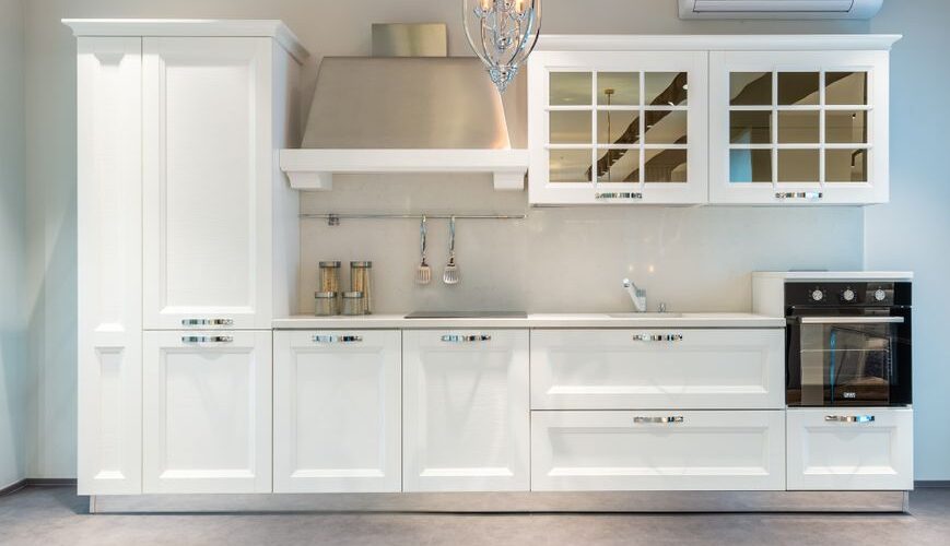 White modern kitchen with tall cabinet, stainless range hood, glass-front upper cabinets, and built-in oven.