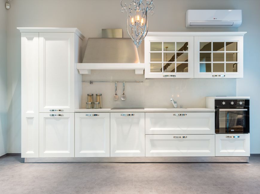 White modern kitchen with tall cabinet, stainless range hood, glass-front upper cabinets, and built-in oven.